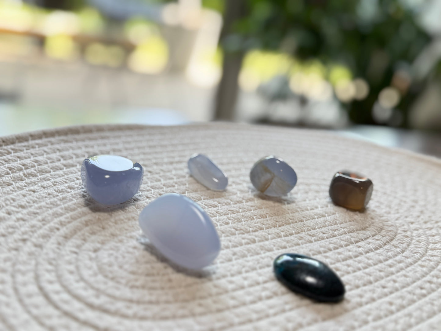 背景のグリーンが柔らかくぼけた構図。手前にブルーカルセドニーの丸みのある石が並び、奥に向かってクリソコラと他の石が静かに並ぶ。