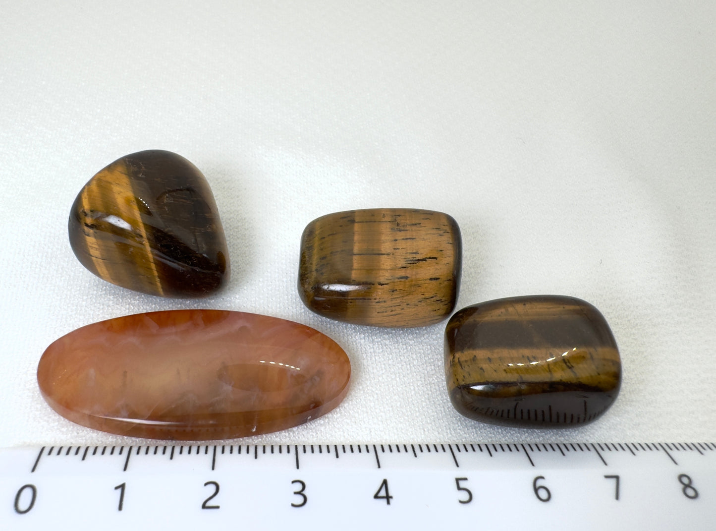 白背景に置かれた天然石4点。中央にカーネリアン(約45mm)が横向きに配置され、その上部に3つのタイガーアイが整然と並ぶ。下部に定規が映り込み、サイズ感が明確に伝わる構図。