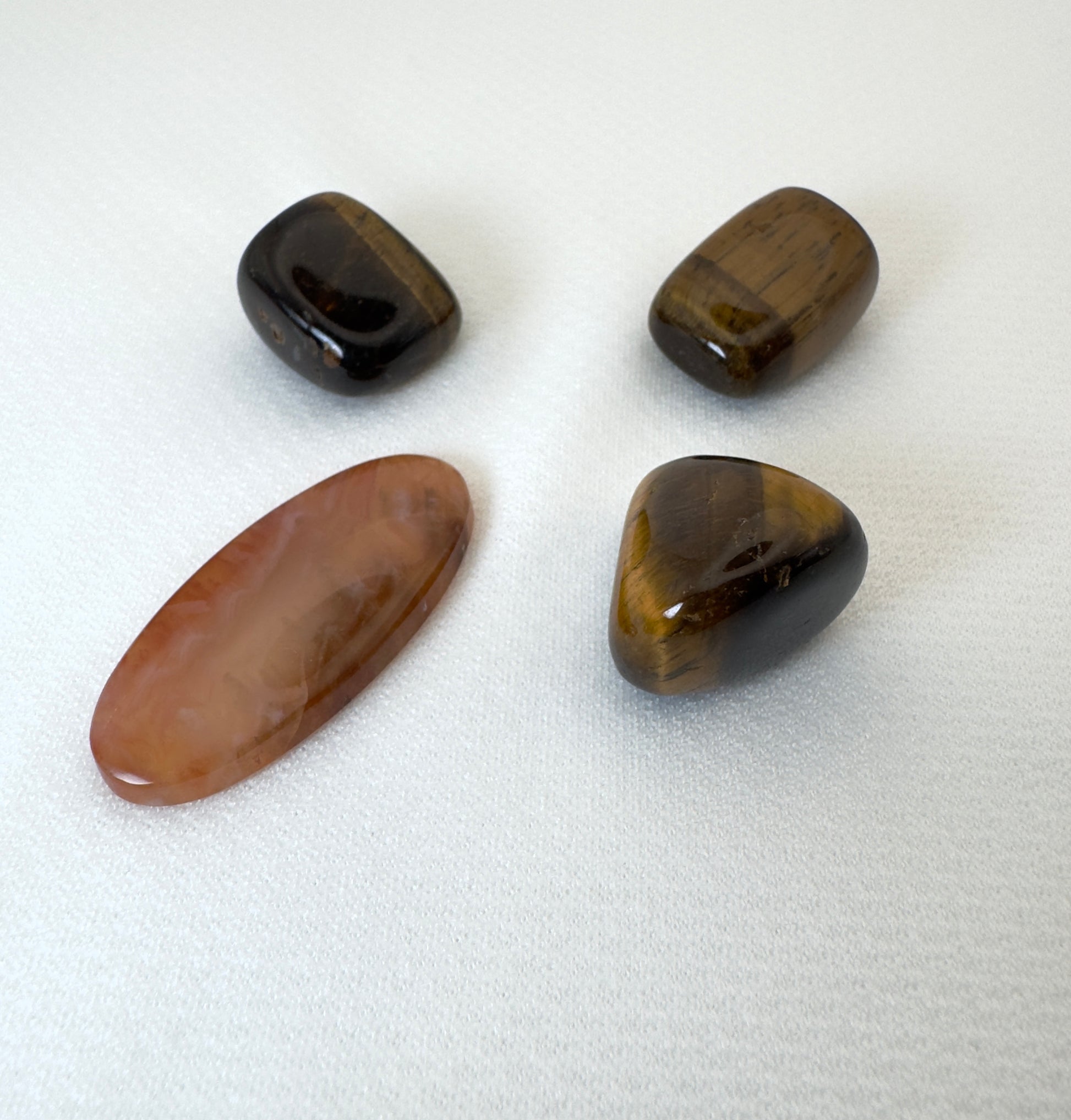 白布の上で斜めに並ぶ4つの天然石。横置きのカーネリアンに、3つのタイガーアイがゆるやかに沿うように配置され、立体感と石の光沢が際立つ。
