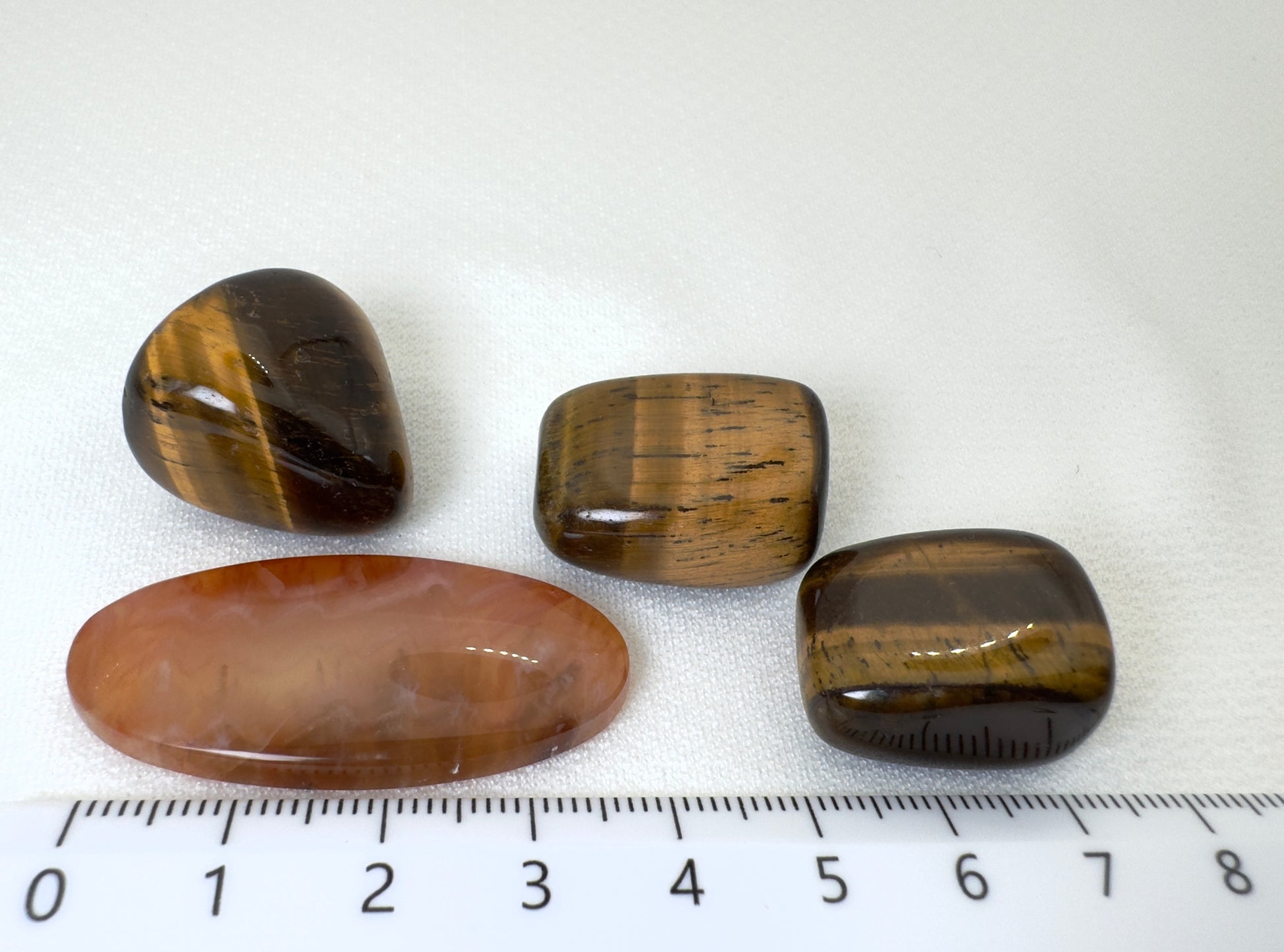 白背景に置かれた天然石4点。中央にカーネリアン（約45mm）が横向きに配置され、その上部に3つのタイガーアイが整然と並ぶ。下部に定規が映り込み、サイズ感が明確に伝わる構図。